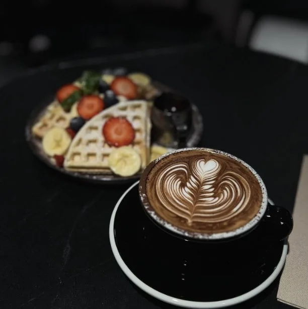 A latte with art in a black cup beside a waffle topped with fruit
