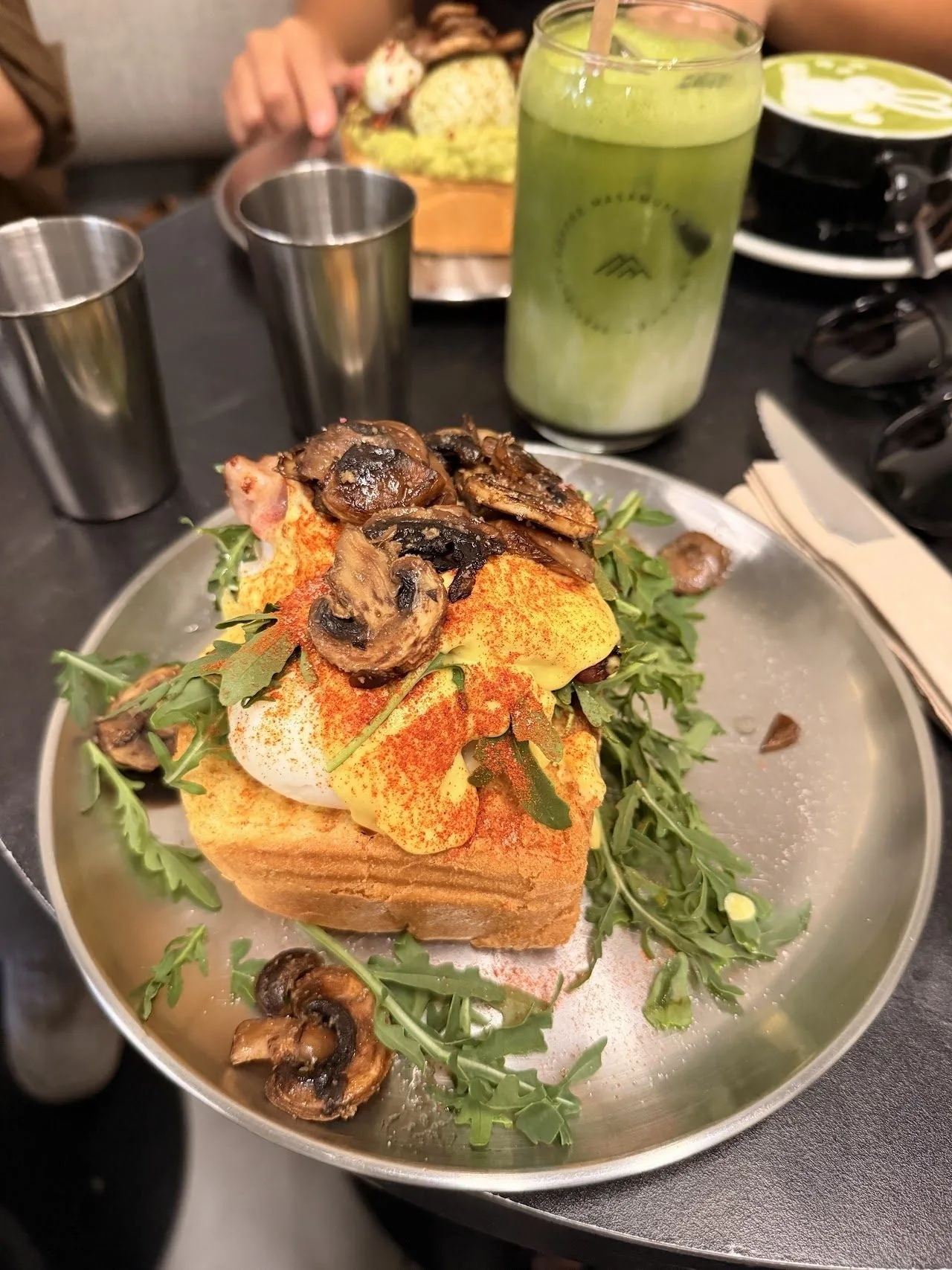 A plate of toast topped with mushrooms and greens beside glasses on a table