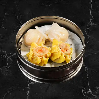 Dim sum in a metal steamer basket - translucent har gow shrimp dumplings and yellow shumai on a dark marble surface