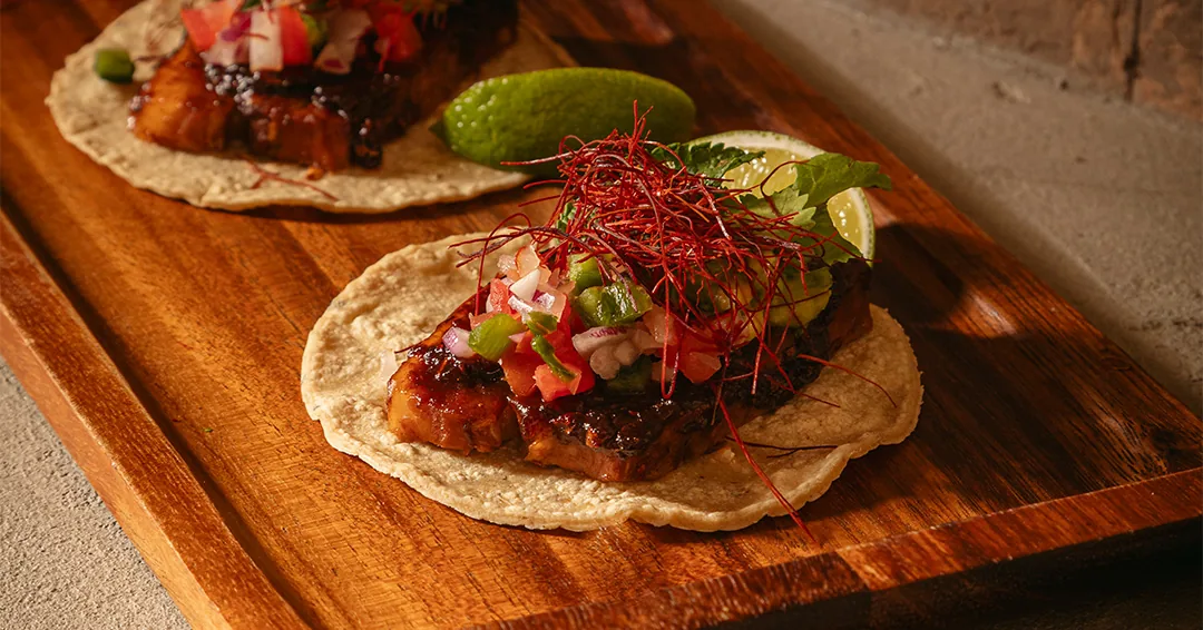 Two soft tacos topped with diced tomato, cilantro, and red garnish on a wooden board