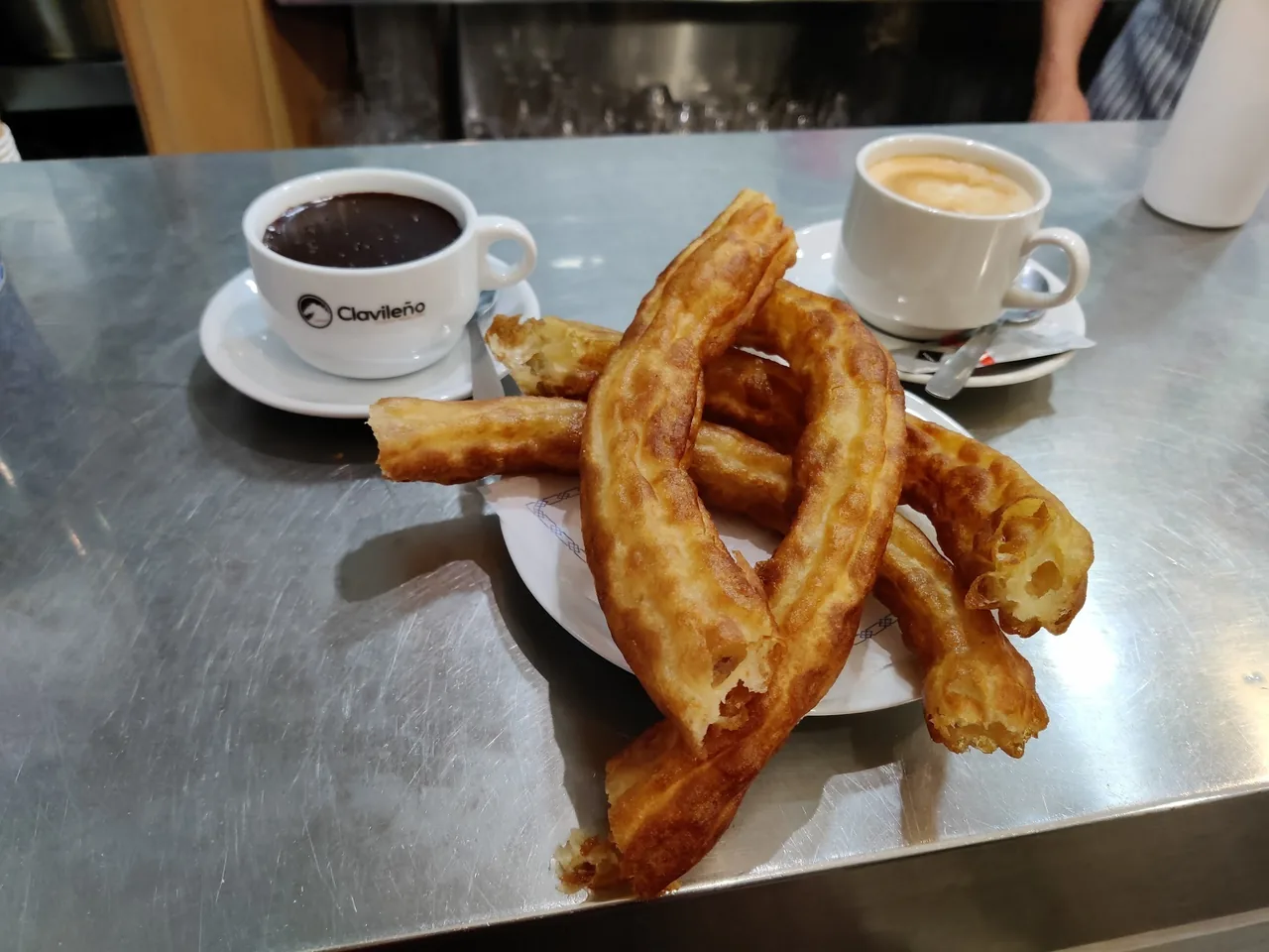 A plate of golden churros beside a cup of hot chocolate and a coffee on a steel counter