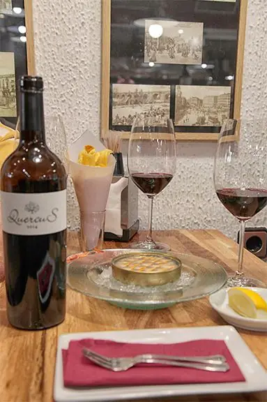 A wooden table set with a bottle of Quercus red wine, two glasses of red wine, a paper cone of crisps, a round dish on a bed of ice and a small dish of lemon wedges, framed sepia photos on the wall behind
