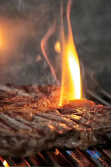 A marbled steak searing over open flames on a grill with smoke rising