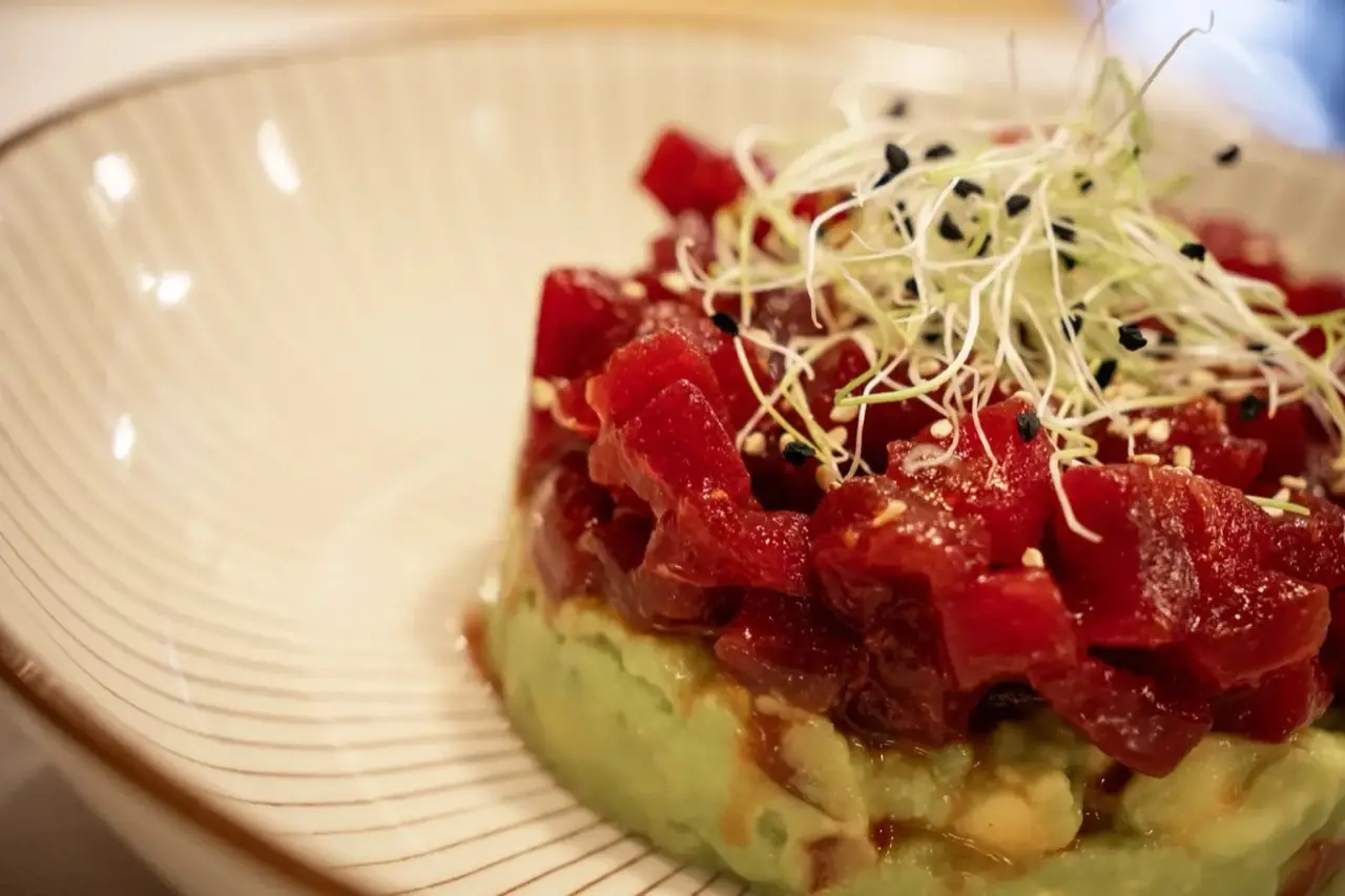 Diced red tuna tartare mounded over mashed avocado in a cream ridged bowl, topped with sprouts and black-and-white sesame seeds