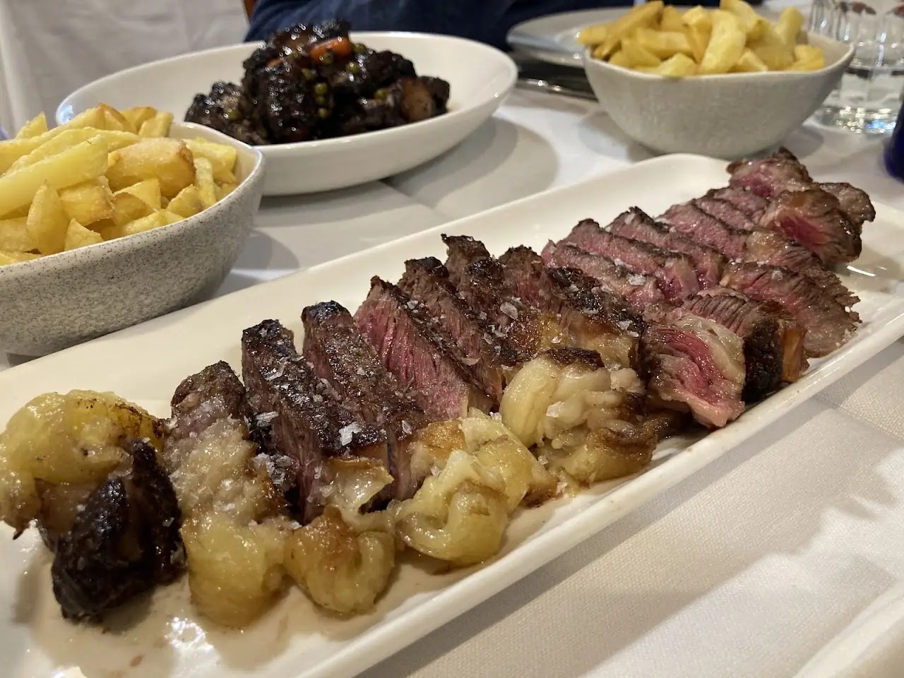 A sliced steak platter with roasted potatoes and a side of fries on a long white plate.