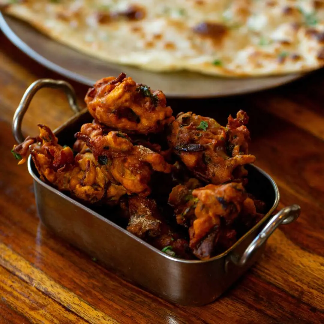Crispy fried onion pakora fritters in a metal dish with naan bread in the background, Mayura
