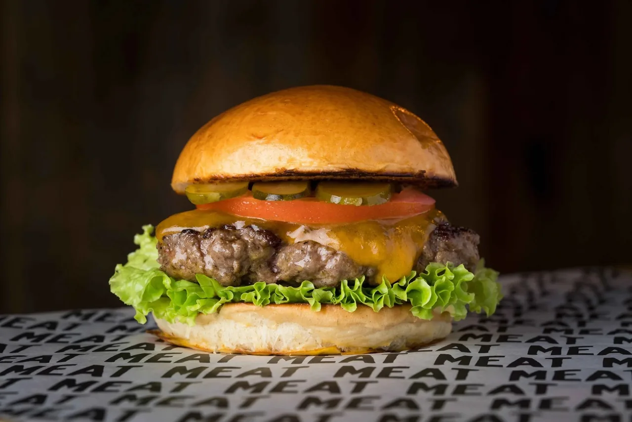 A cheeseburger with lettuce, tomato and pickles on a brioche bun, set on printed paper against a dark background