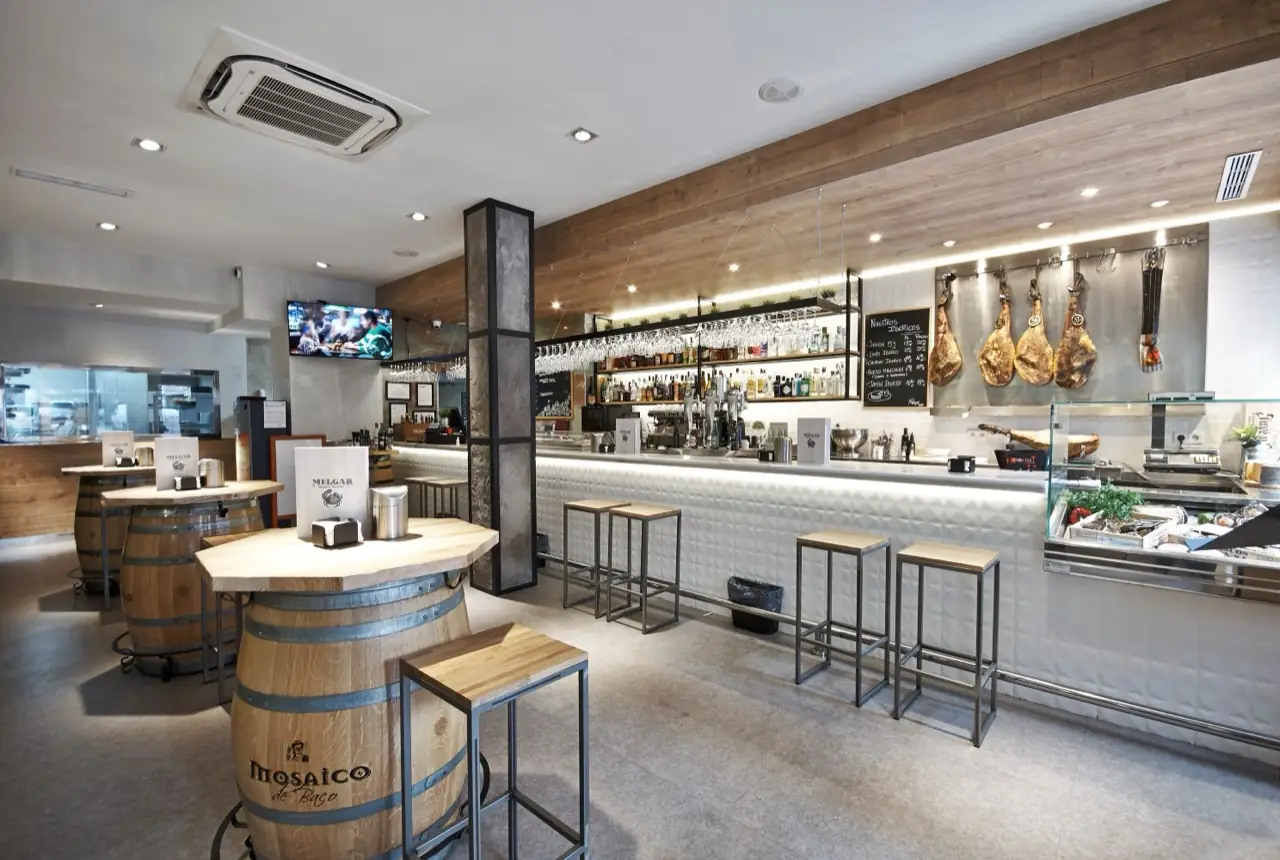 Interior of Melgar bar with barrel high-tables and stools, a long white-tiled bar backed by bottles, hanging cured hams and a seafood display case