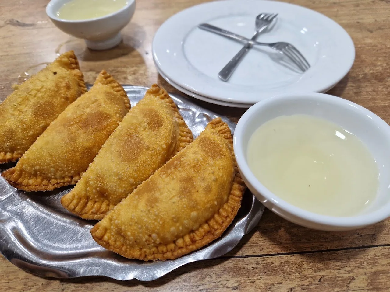 Three golden fried turnovers on a metal plate beside a small dish of sauce