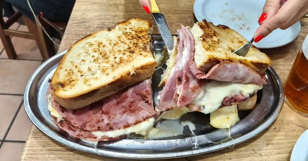 A large toasted sandwich of ham and melted cheese on a metal oval tray, being cut with a knife and fork