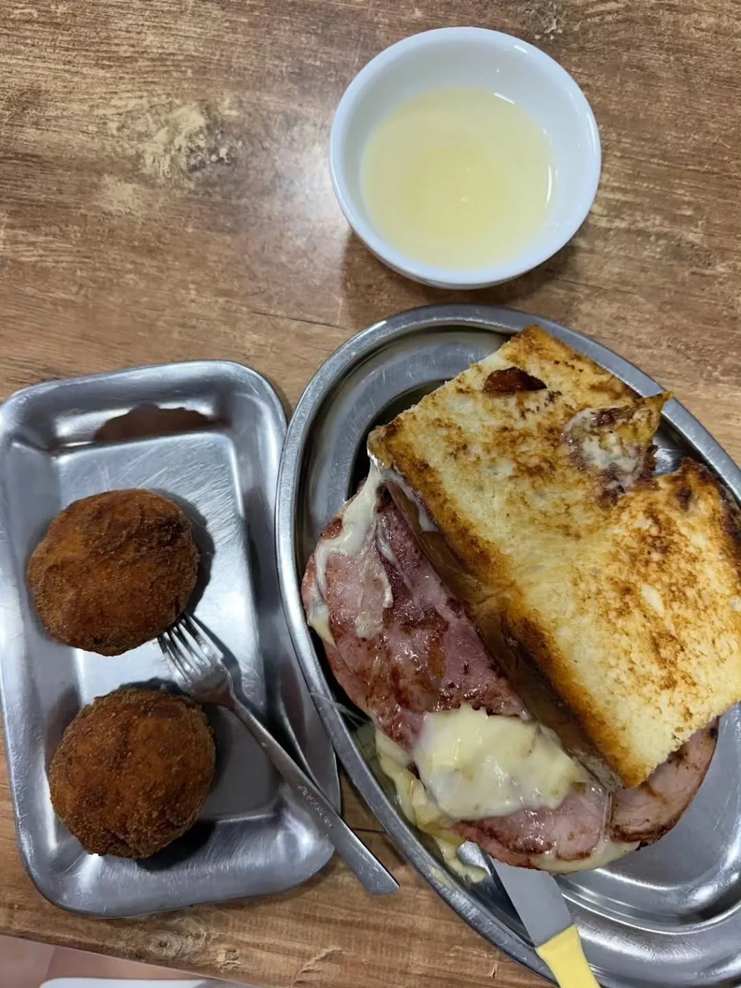 A toasted ham and cheese sandwich on a metal oval tray beside two croquettes on a wooden table
