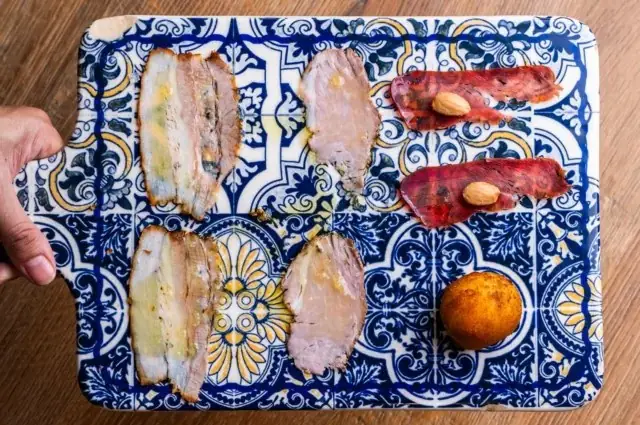 Slices of terrine and cured meat with almonds and a croquette arranged on a blue-and-yellow decorative tile board held by a hand