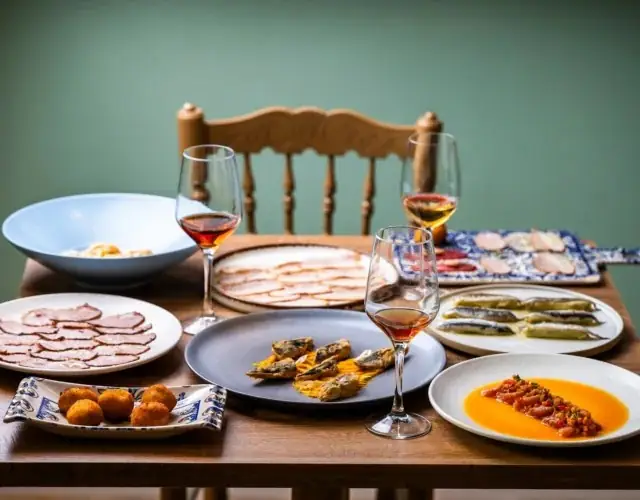 Wooden table set with several sharing plates - cured meats, croquettes, fried fish, marinated anchovies and a tomato dish - alongside glasses of amber wine against a green wall