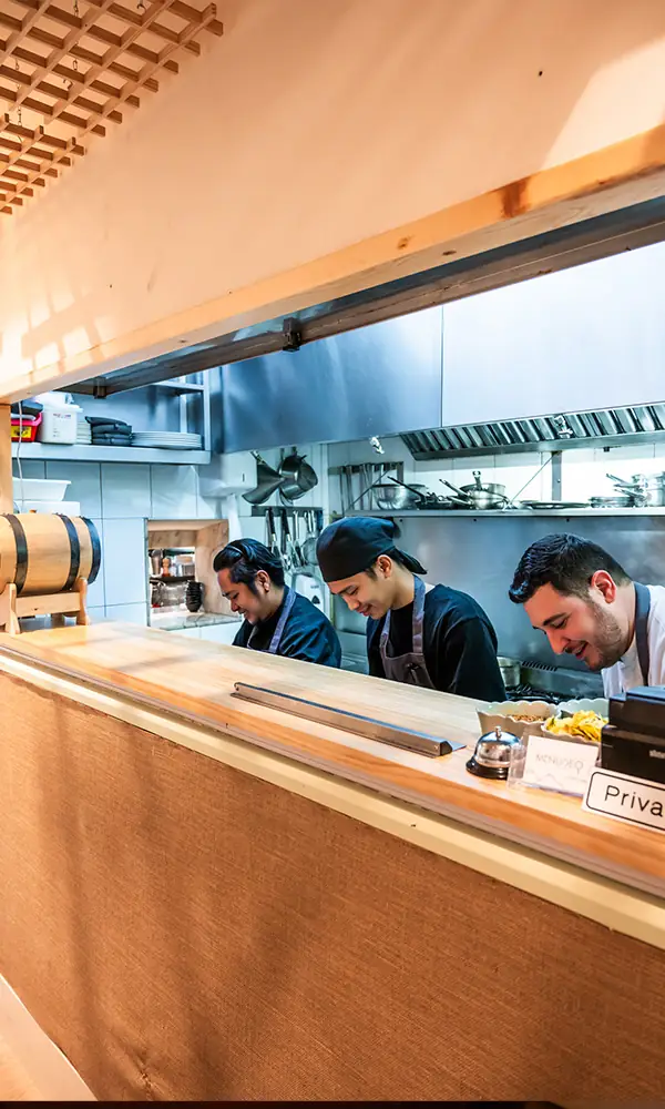 Open kitchen pass seen over a light-wood bar counter, three cooks in dark aprons working at the stainless-steel line, a wooden barrel to the left