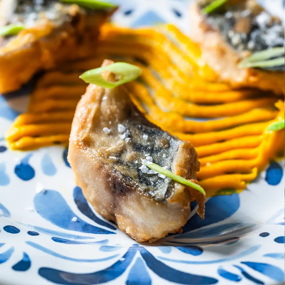 Pieces of crisp-fried mackerel topped with spring onion and flaked salt over a piped orange puree on a blue-and-white patterned plate