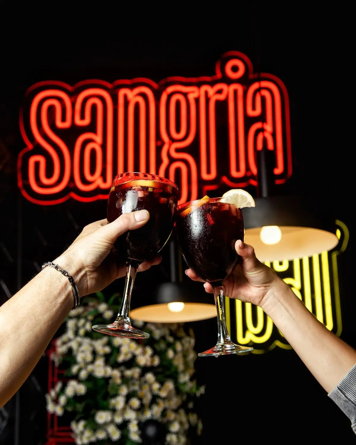 Two hands toasting balloon glasses of red sangria in front of a red neon Sangria sign