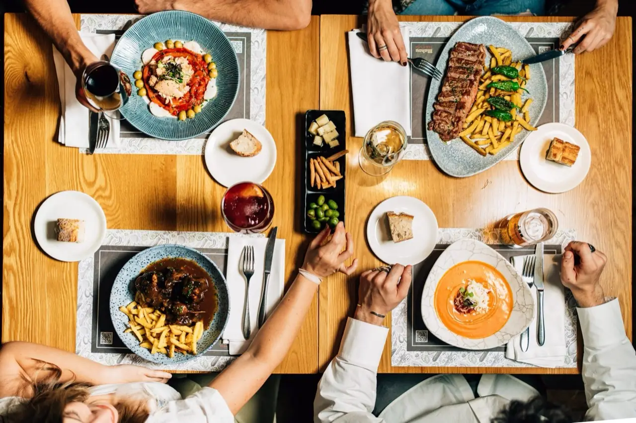 Overhead flat-lay of a wooden table spread with steak and fries, oxtail stew, salmorejo, tomato salad, bread, olives and drinks
