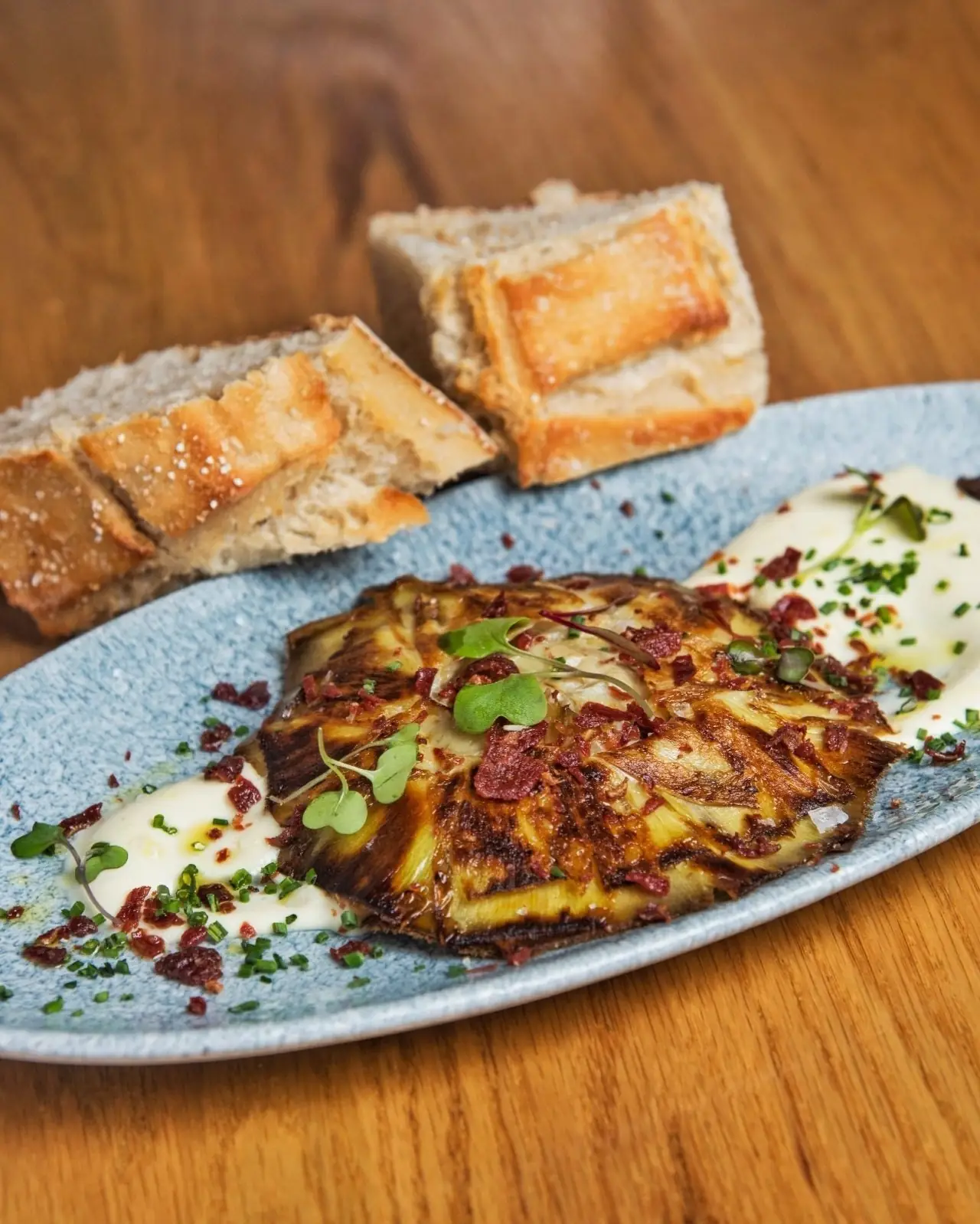 Charred grilled artichoke topped with crispy jamon and microgreens over cream, with crusty bread on a blue plate