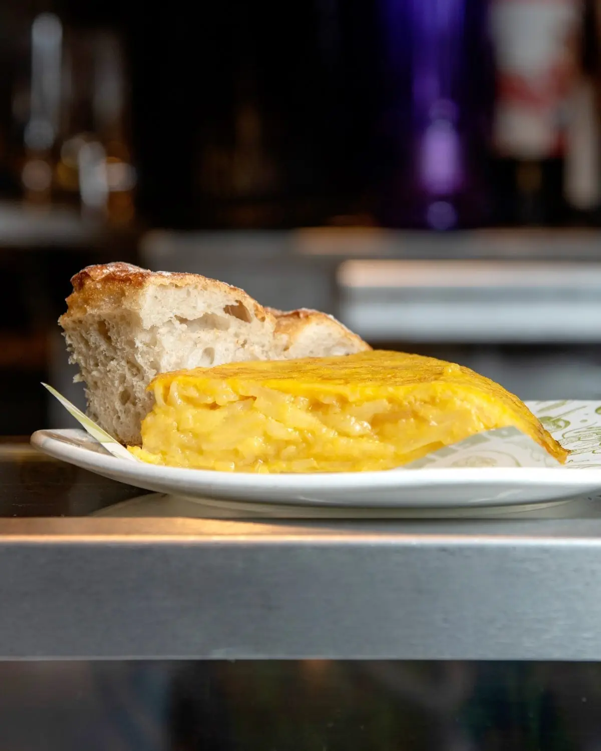 Slice of runny Spanish tortilla with a chunk of rustic bread on a white plate at a bar counter