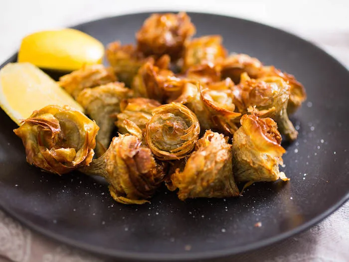 Fried whole artichokes on a black plate with lemon wedges