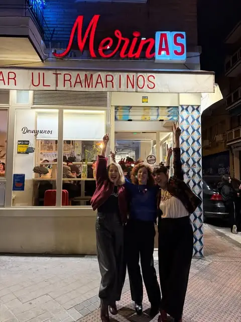 Night exterior of Merinas, a bar with red neon sign reading Merinas and BAR ULTRAMARINOS above a white storefront with awning