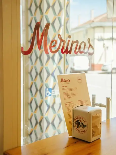 Window table detail with the Merinas name painted in red script on the glass, a paper menu propped up and a Victoria Malaga napkin dispenser on a wooden ledge
