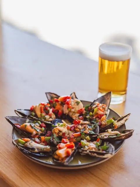 Plate of mussels dressed with diced red and green peppers in vinaigrette beside a glass of golden beer on a wooden table