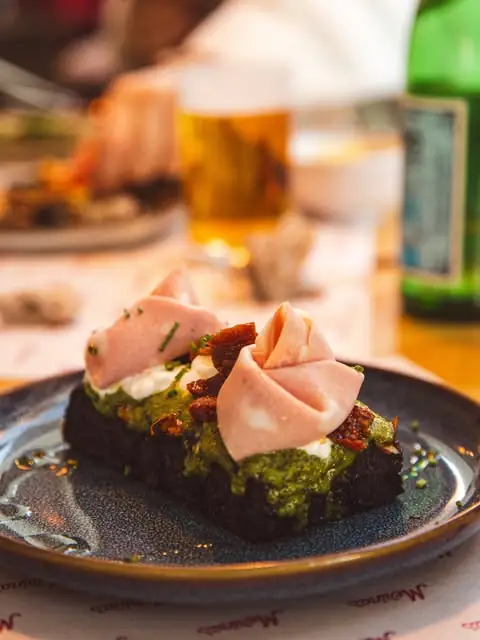 Open-faced toast on dark bread topped with green pesto, sundried tomato and rosettes of mortadella, on a dark plate with a beer behind
