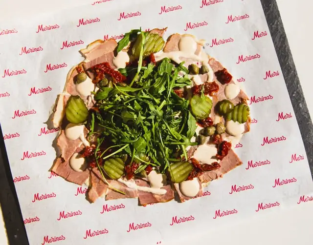 Overhead plate of thin-sliced cooked ham topped with arugula, capers, gherkins, sundried tomato and creamy dressing, on Merinas-branded paper