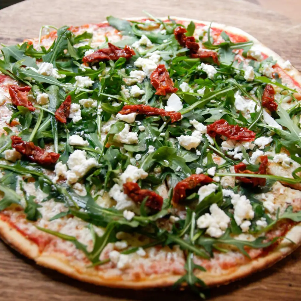 Gluten-free pizza topped with arugula, crumbled goat cheese, and sun-dried tomatoes on a wooden board at Messié Pizza Gluten Free Gràcia.