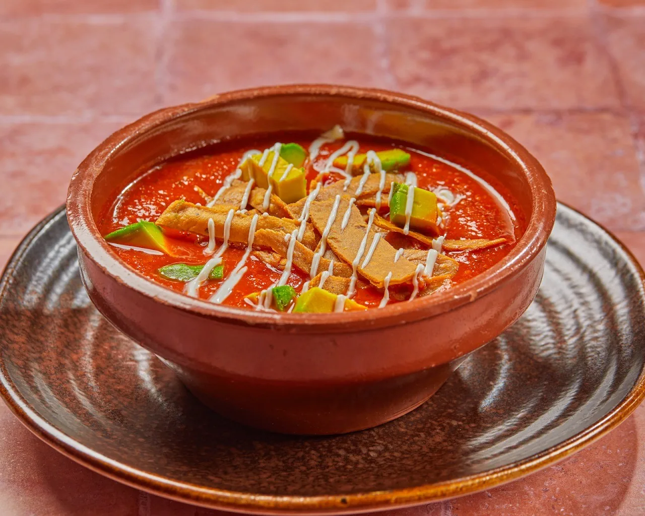 A terracotta bowl of red broth topped with avocado slices and cream