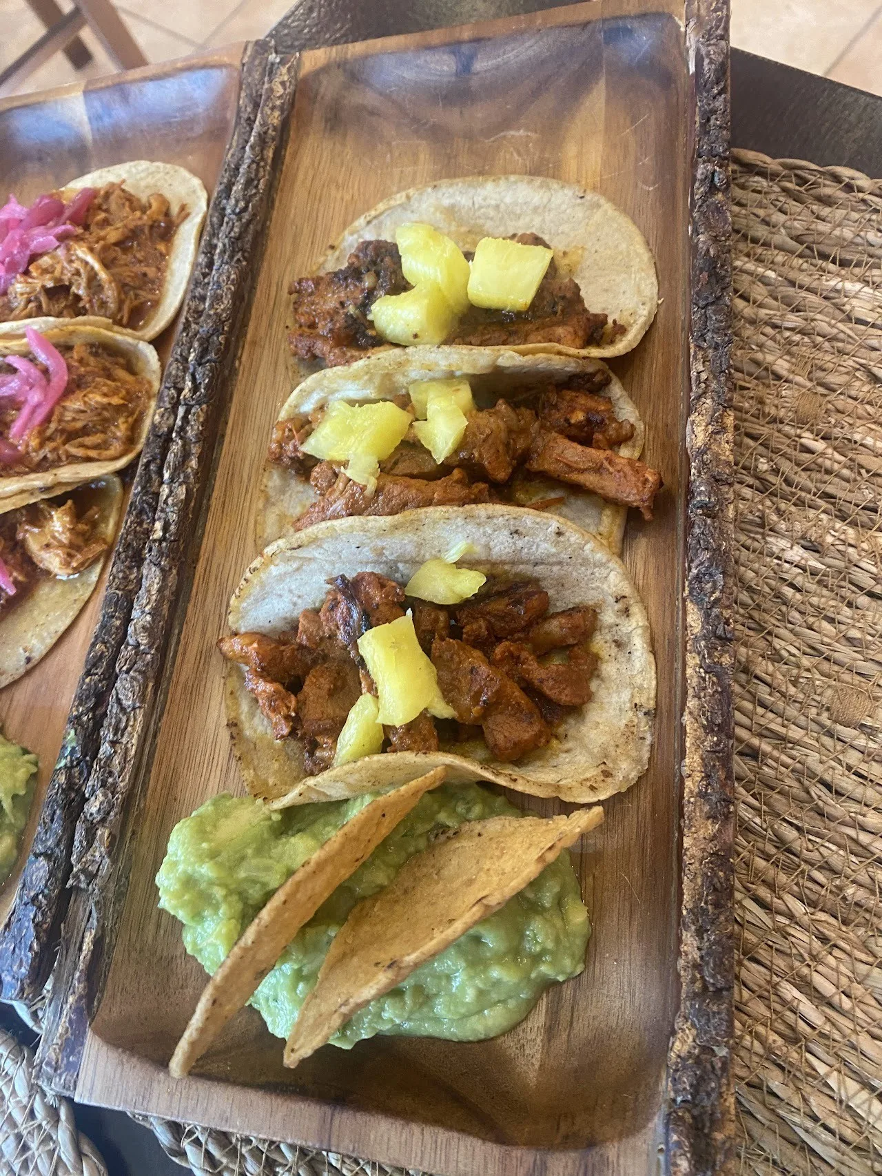 A wooden board of tacos filled with sliced meat and pineapple, with guacamole underneath
