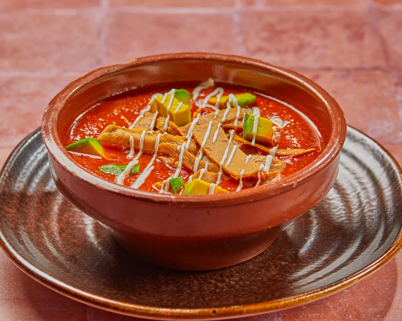 A bowl of red pozole-style soup garnished with avocado and a drizzle
