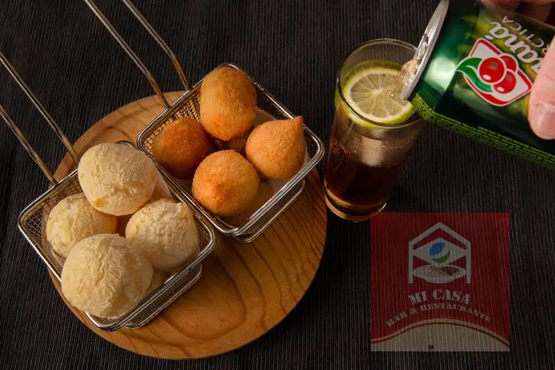 Pão de queijo and coxinha in metal fry baskets on a wooden board, with a glass of guaraná soda over ice with lime, Mi Casa Bar y Restaurante