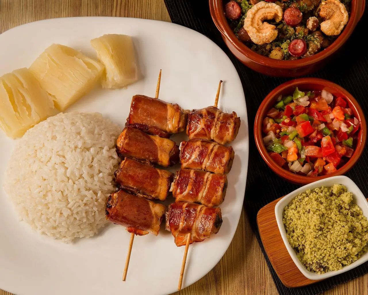 Bacon-wrapped meat skewers on a plate with white rice and boiled cassava, alongside small bowls of farofa and tomato-pepper salsa, Mi Casa Bar y Restaurante