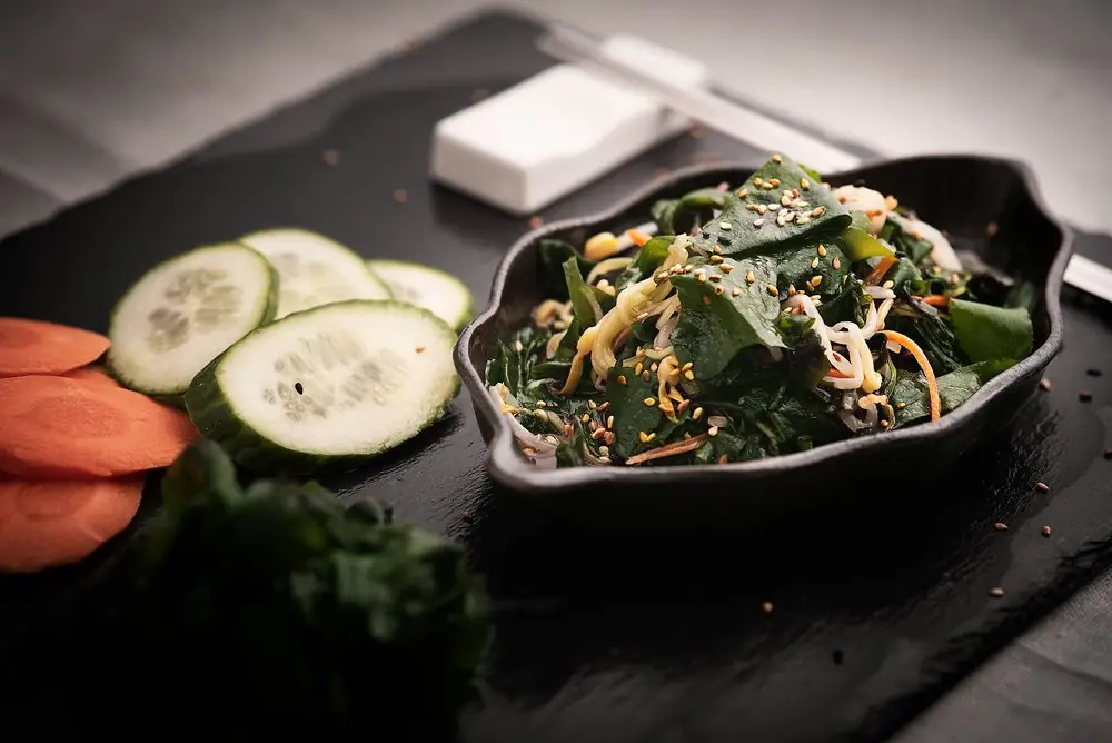 Seaweed salad with bean sprouts, carrot and sesame seeds in a black dish, with sliced cucumber and carrot alongside on slate