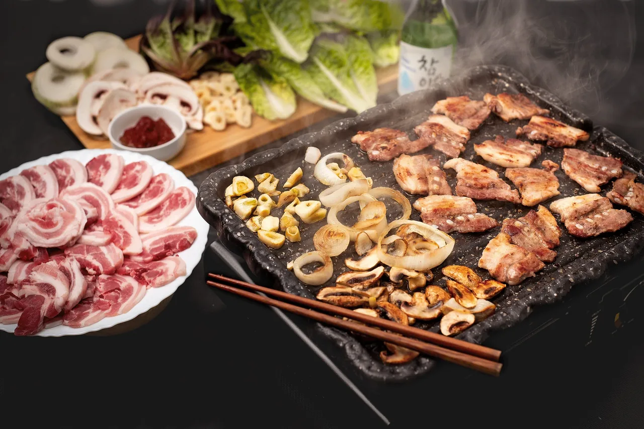 Sliced pork belly grilling on a black stone plate with garlic, onion and mushrooms, with lettuce and a bottle behind