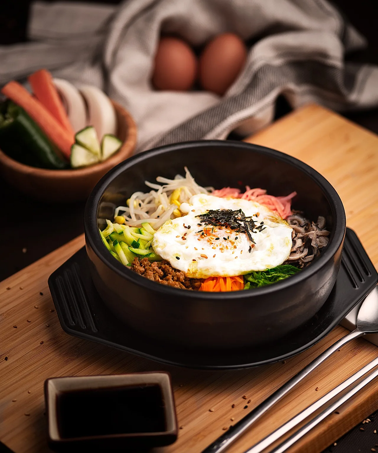 A black stone bowl of rice topped with a fried egg, sprouts and vegetables on a wooden board