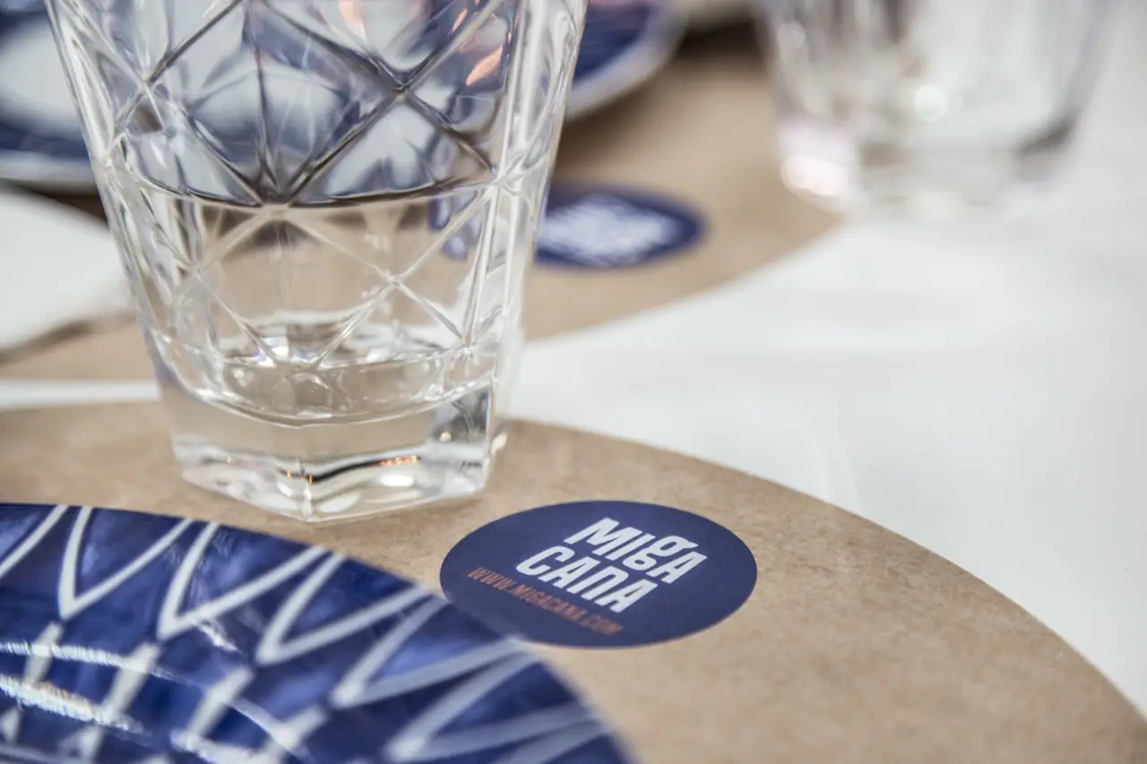 Close-up table setting with a cut-glass tumbler of water on kraft-paper placemat bearing the round Miga Cana coaster and a blue patterned plate