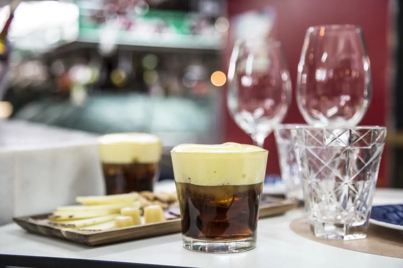 Tumbler of dark vermouth with a frothy foam head beside a wooden board of sliced cheese and wine glasses on a marble bar