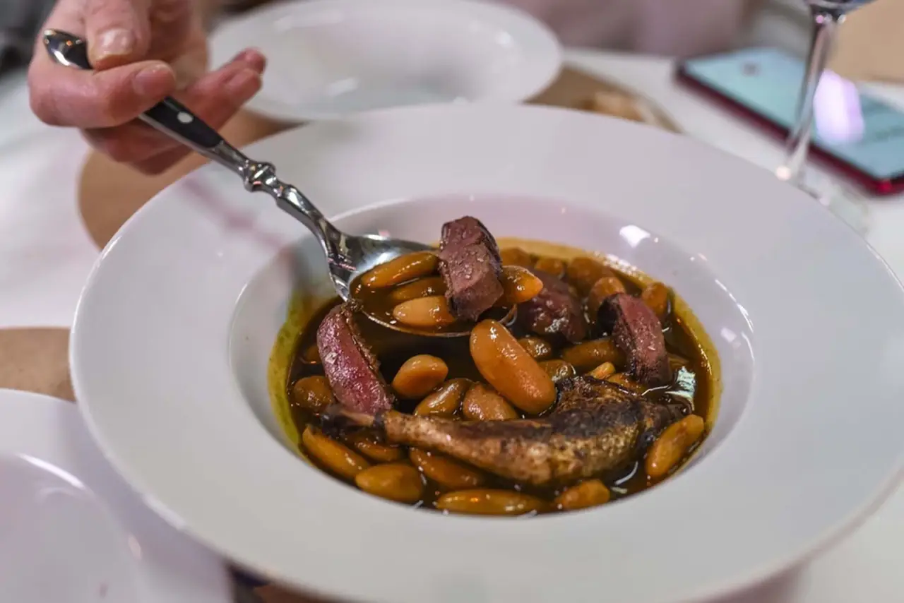 White bowl of white-bean and game-bird stew, a spoon lifting a piece of dark meat from the broth