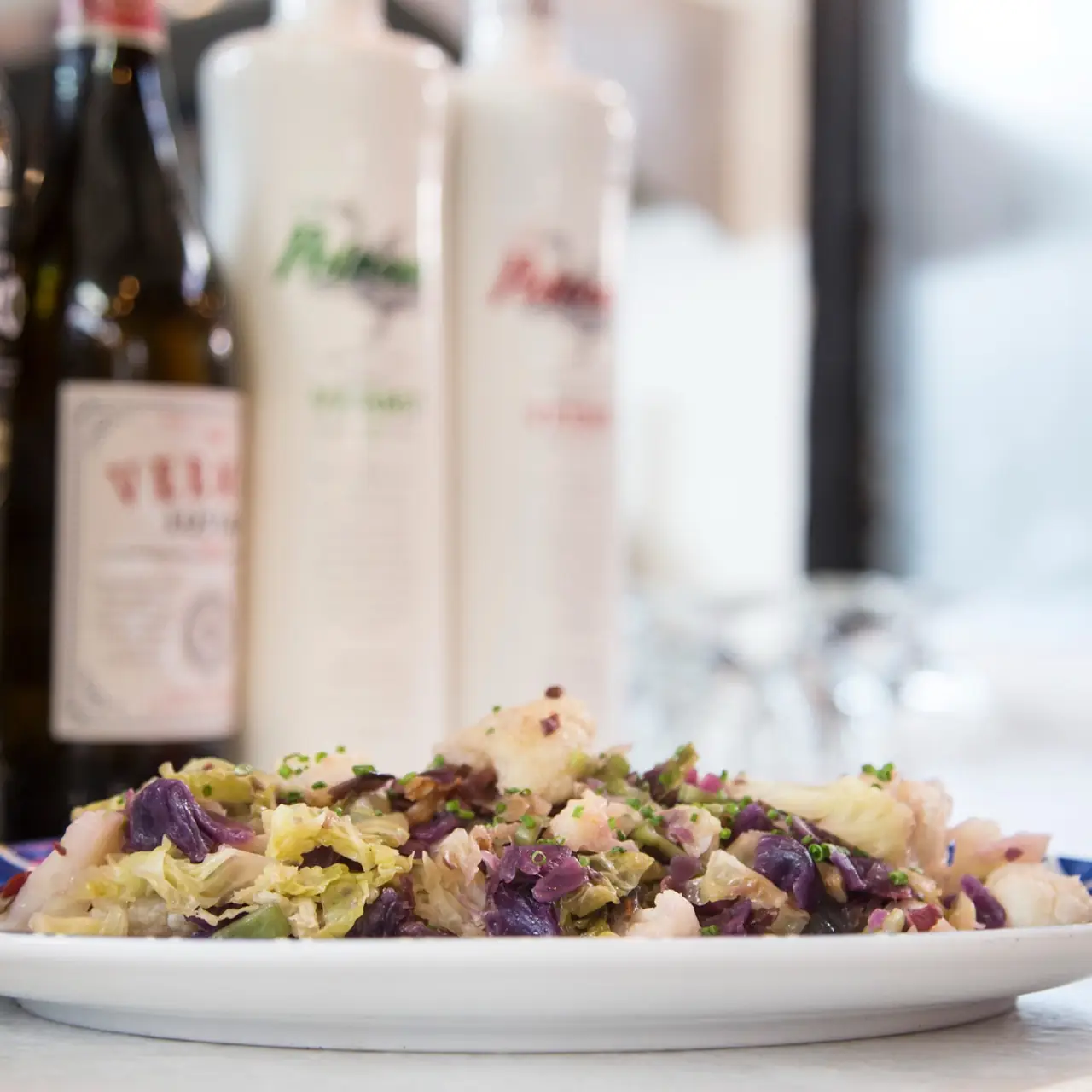 Plate of sauteed green and red cabbage with cauliflower and chives, vermouth and condiment bottles behind