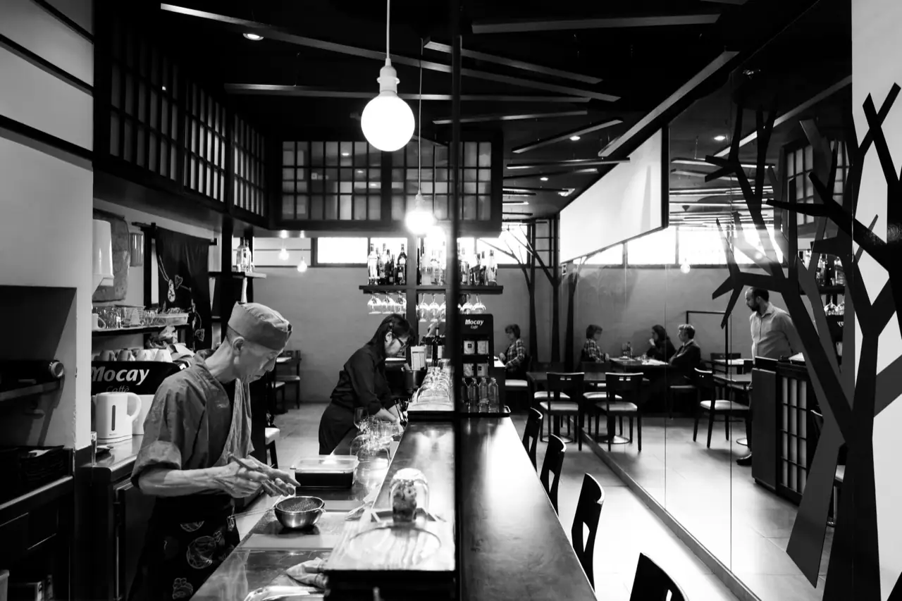 Black-and-white interior of a Japanese restaurant with a long sushi counter, a chef in uniform working, hanging bulbs and diners at tables behind glass