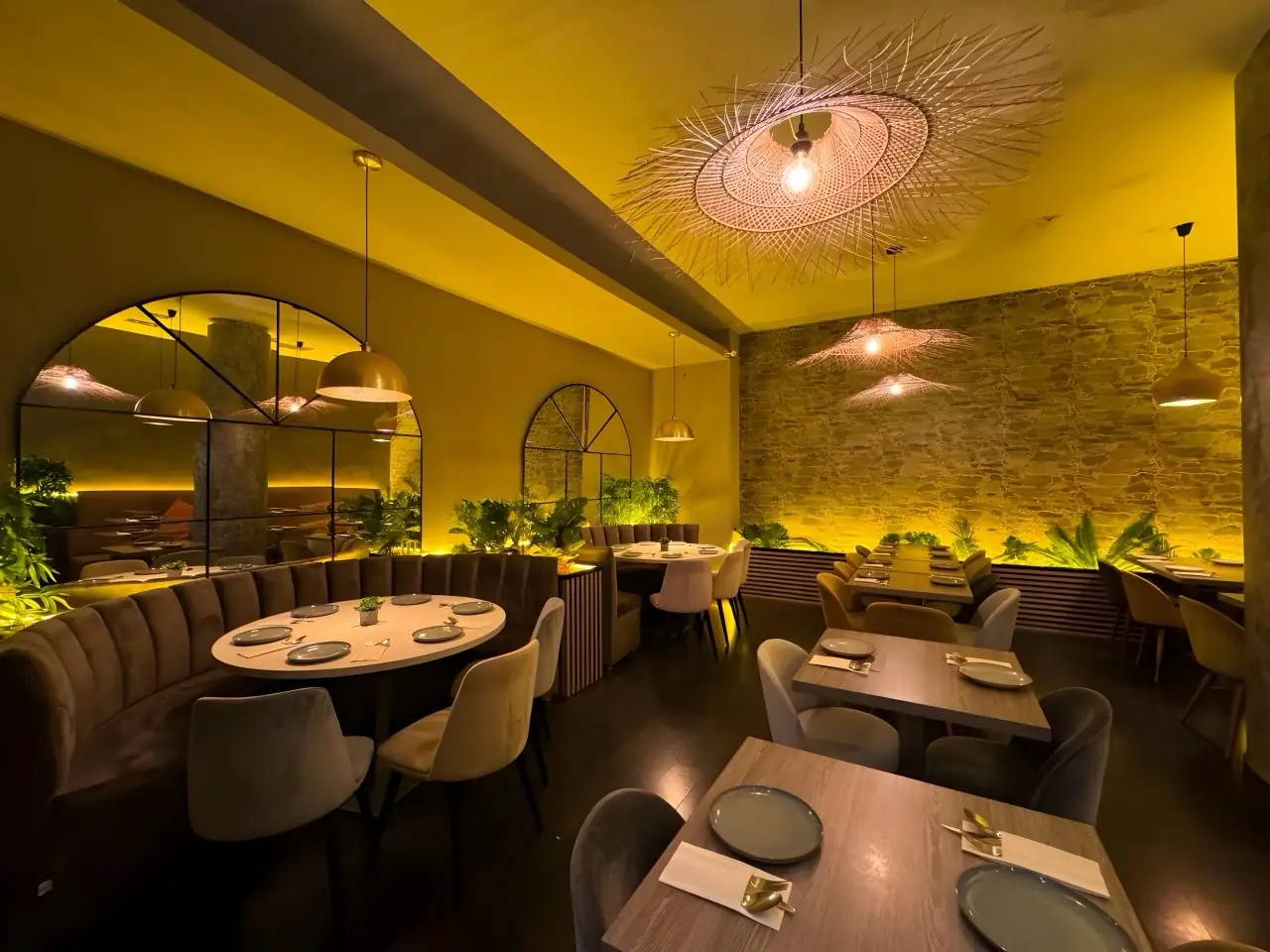 Warm-lit dining room with olive-green walls, velvet banquette around an oval table, rattan pendant lamps and a plant-covered stone wall