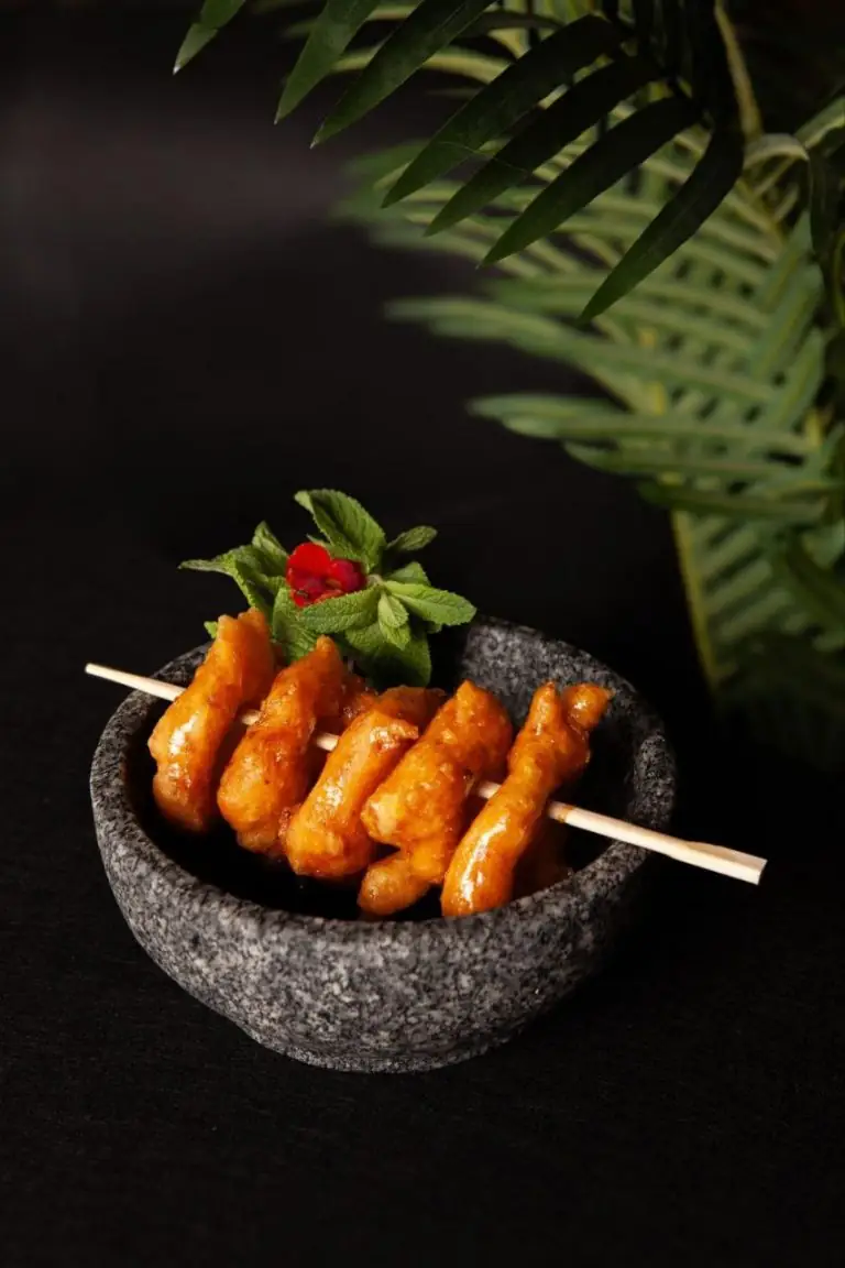 Skewer of glazed fried chicken pieces topped with mint in a small grey stone bowl on a black surface