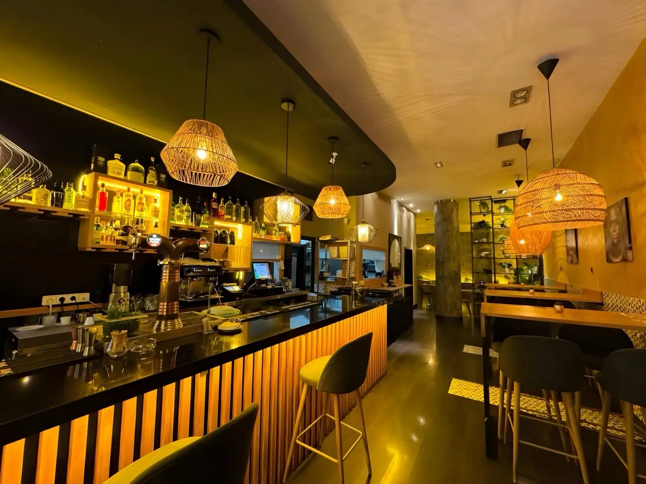 Dimly lit bar counter with wooden slat front, backlit liquor shelves, espresso machine and woven pendant lamps