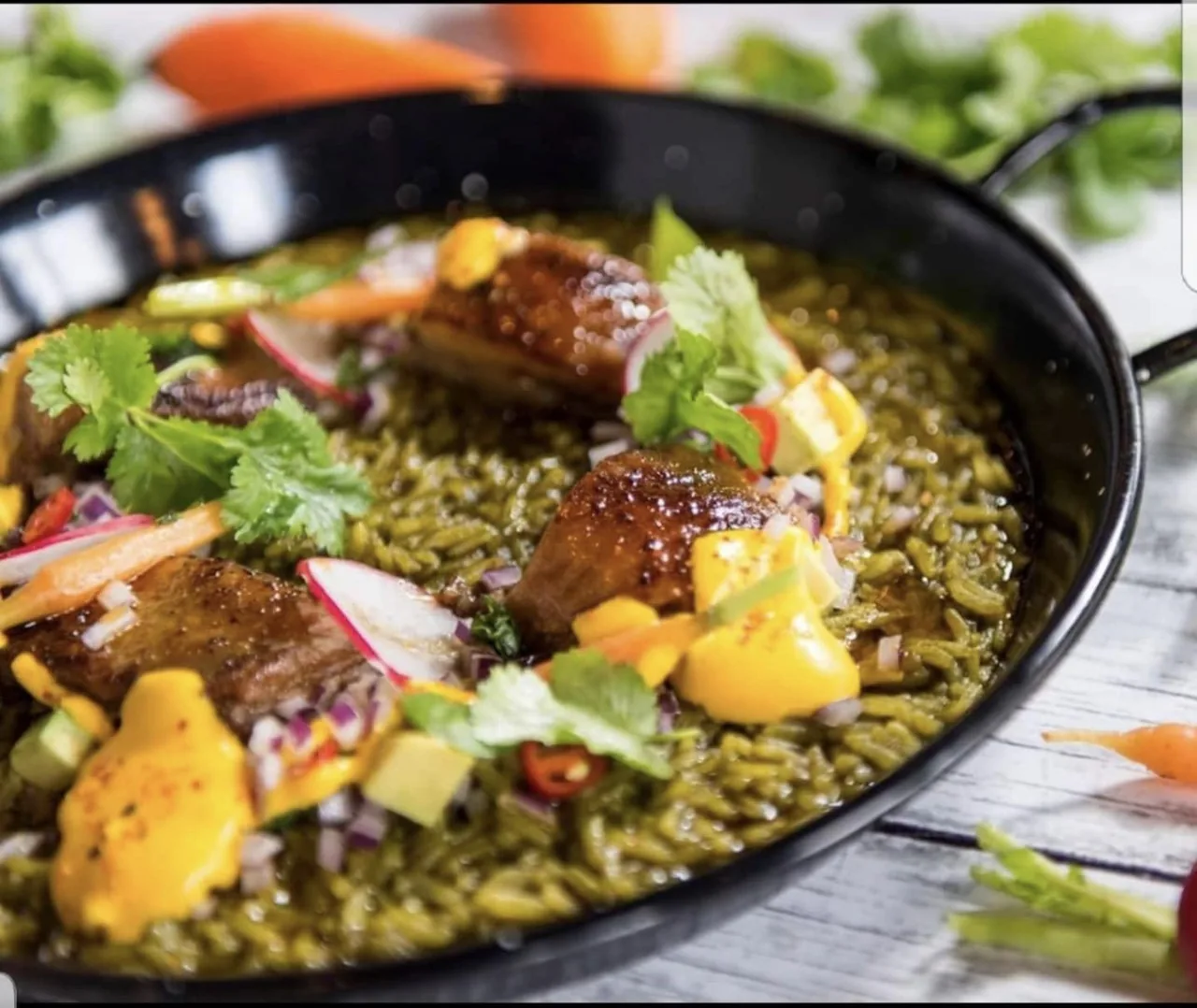 Green rice with pieces of glazed meat, vegetables and yellow sauce in a black pan
