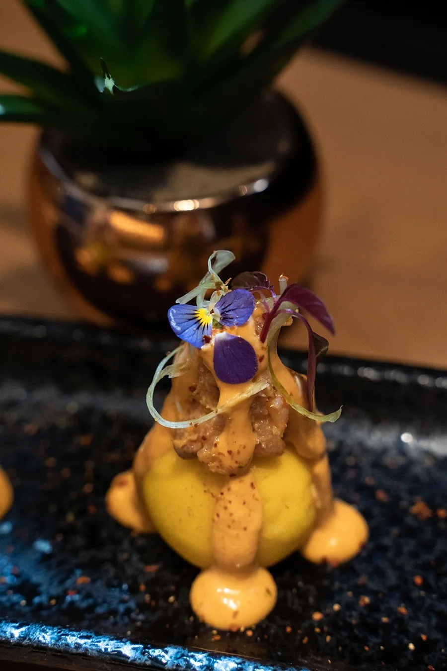 A round yellow potato dish topped with a pink sauce and edible flowers on a dark speckled plate