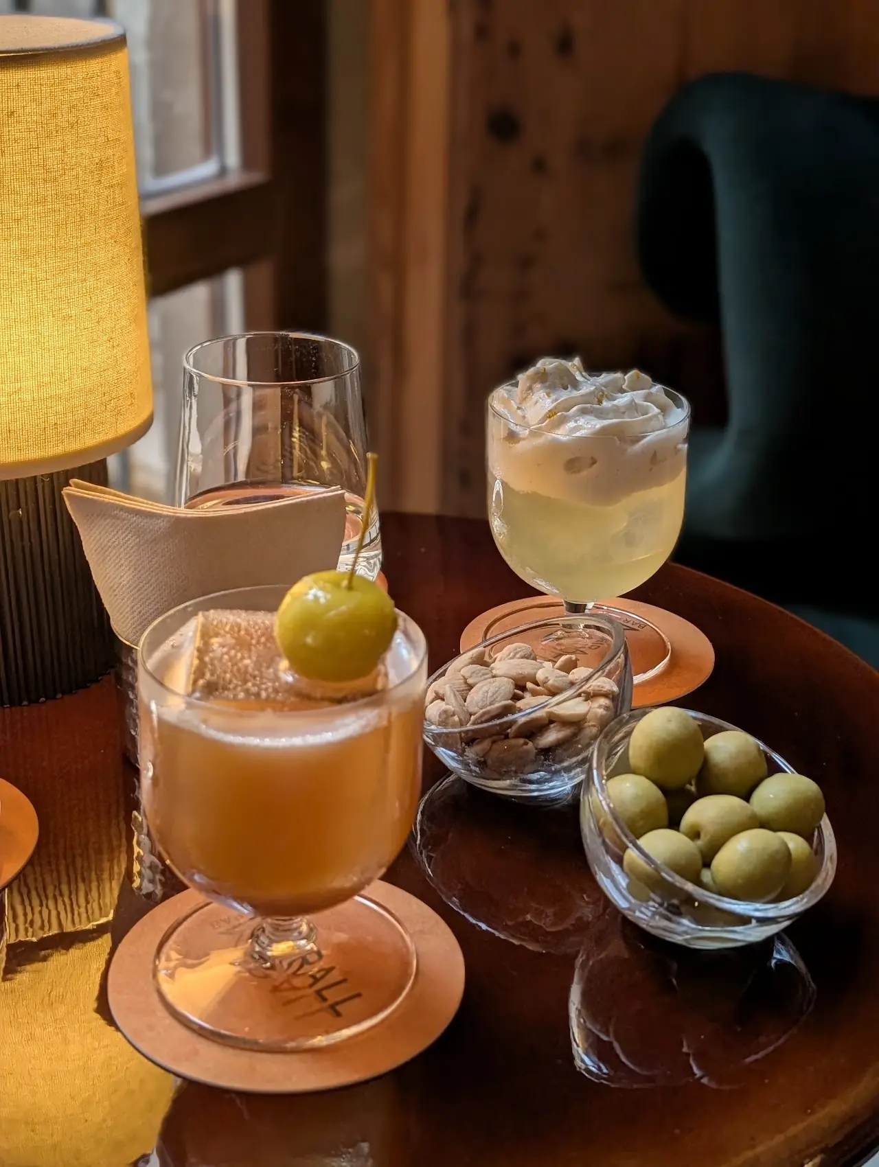 Two cocktails, one garnished with an olive, next to small bowls of olives and pistachios on a bar table.
