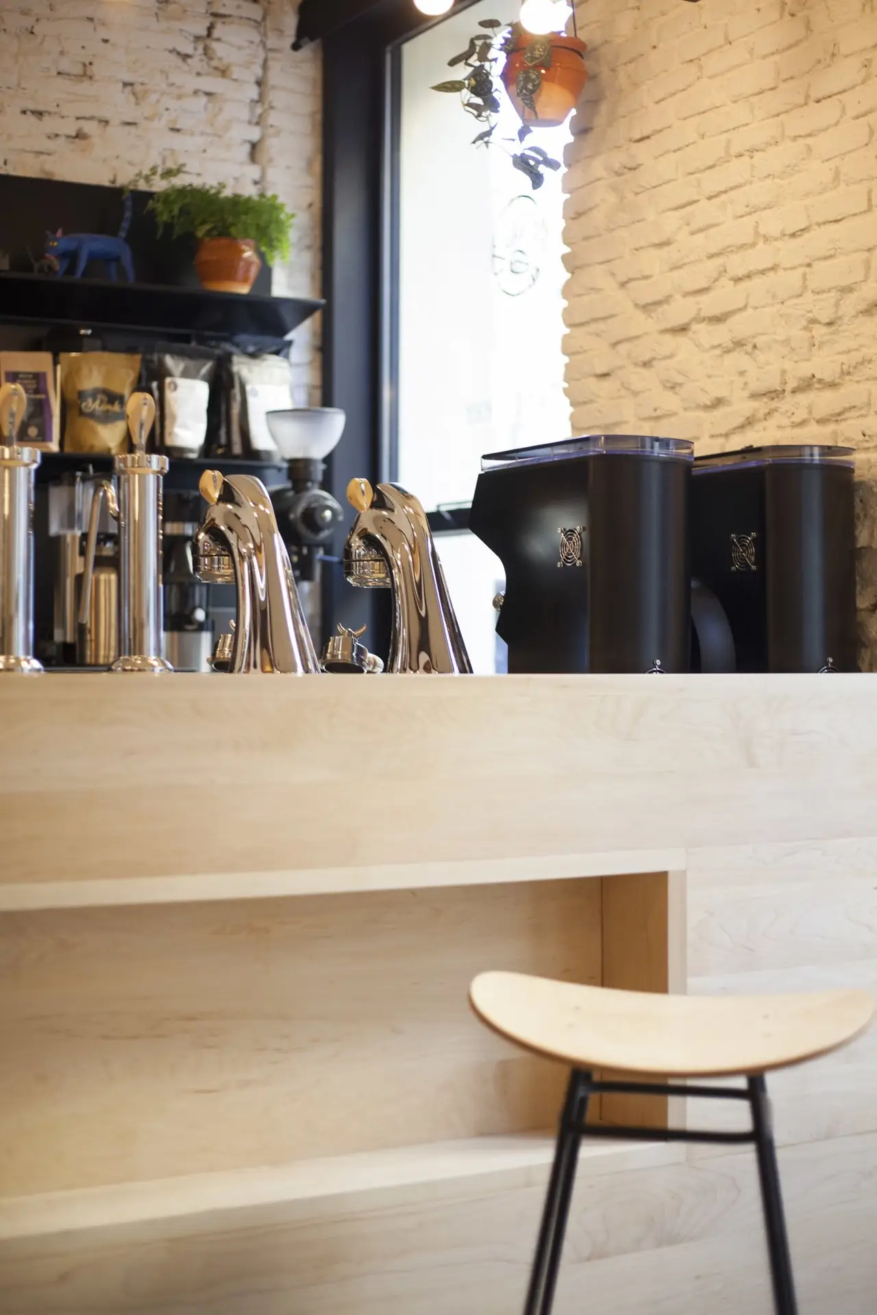 Cafe bar counter with chrome espresso machine group heads, a coffee grinder and shelves of retail coffee bags against exposed brick